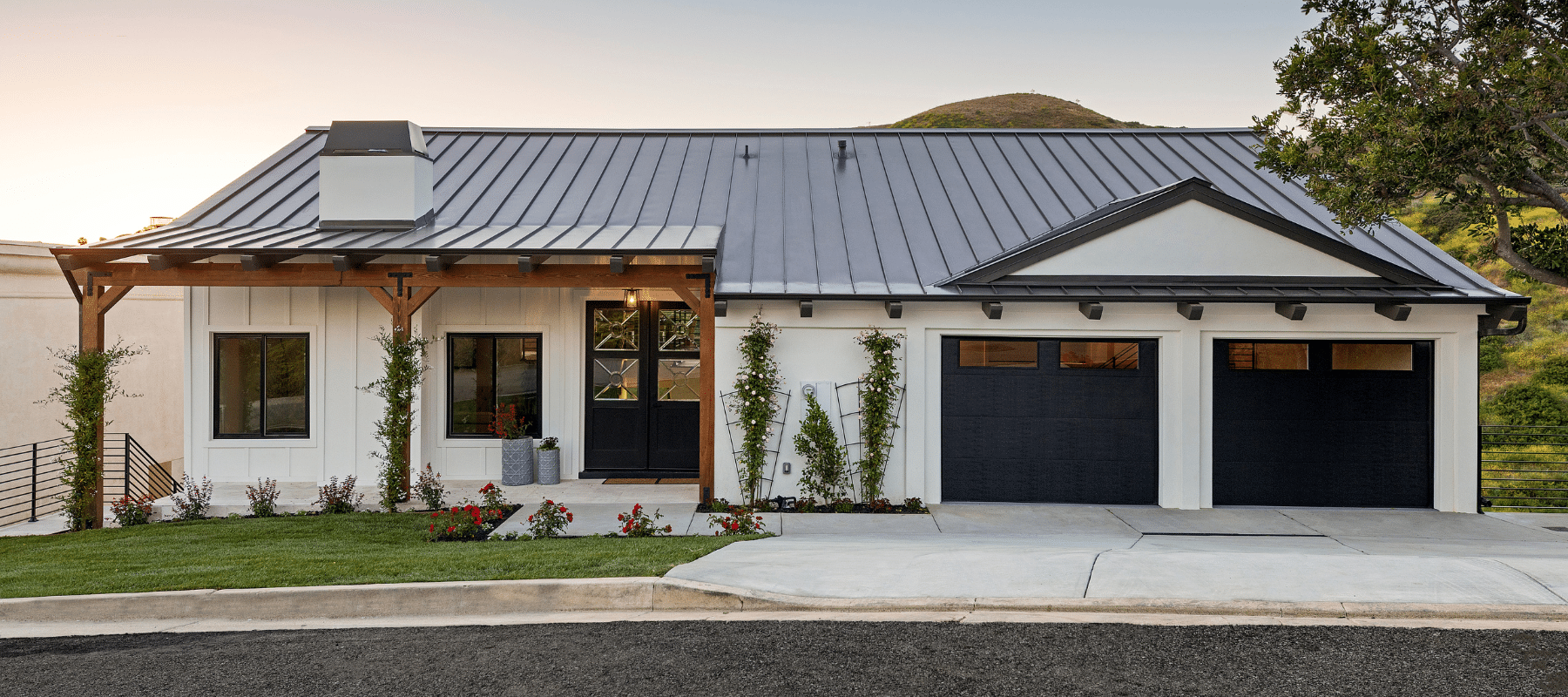 A contemporary white farmhouse with a metal roof and black garage doors at sunset